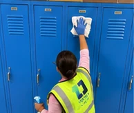 Crew member wiping down blue school lockers, with classroom cleaning in progress